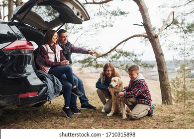 Happy Family Sitting And Having Fun With Their Dog Near Modern Car Outdoors In Forest.