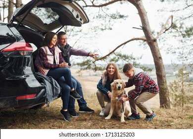 Happy Family Sitting And Having Fun With Their Dog Near Modern Car Outdoors In Forest.