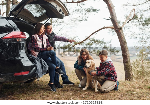 Happy family sitting and having fun\
with their dog near modern car outdoors in\
forest.