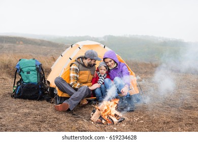 Happy family sitting near tent and looking at burning fire at cloudy autumn day - Shutterstock ID 619234628
