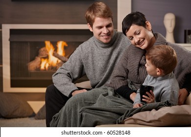 Happy Family Sitting On Couch At Home In Front Of Fireplace, Smiling.