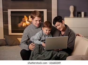 Happy Family Sitting On Couch At Home In Winter, Using Laptop Computer, Smiling.