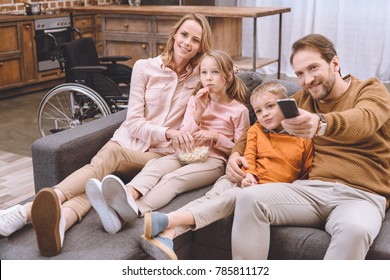 Happy Family Sitting On Sofa And Watching Tv Together And Wheelchair Standing Behind