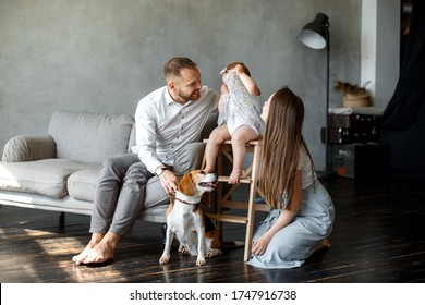 A Happy Family Is Sitting In A Room On The Couch. Father And Mother Are Smiling, The Child Is Shy And Hiding Behind His Dress. Nearby Is A Beagle Dog. Happy Family Concept