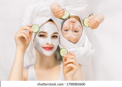 Happy Family Spa Procedures, Mothers Day. Pretty Young Mother And Baby Girl Toddler Lying Head To Head On Isolated White Background, With Face Skin Mask And Towels On Head, Holding Slices Of Cucumber