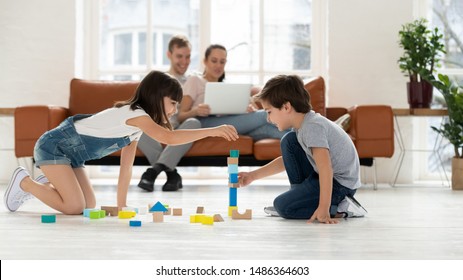 Happy family spend leisure time in living room young parents relax on sofa with laptop, two kids children playing with wooden blocks on warm floor in comfy modern living room with underfloor heating