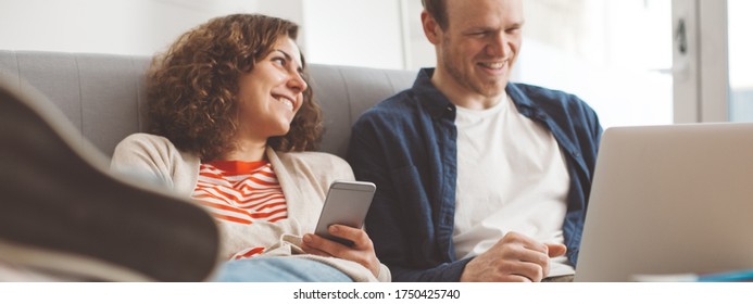 Happy Family Spending Time With Electronic Devices. Man Using Laptop And Smiling Woman Typing Message With Mobile Phone. Sitting Together On The Sofa. Wide Screen Panoramic