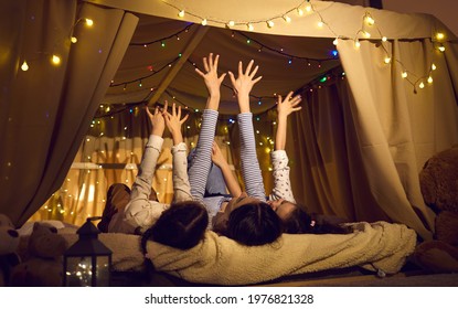 Happy family spending time at home, enjoying quiet evening and having fun together. Mother and little children put hands in air while lying in a cosy bed tent decorated with colourful LED fairy lights