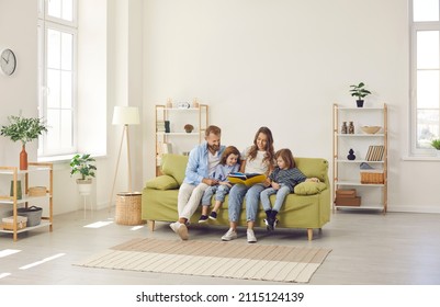 Happy Family Spending Time At Home. Mum, Dad And Little Kids Reading Book While Sitting Together On Green Sofa In Room With Light Walls, Beige Rug And Simple Brown Shelves In Their Big New House