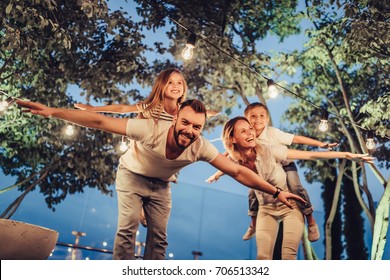 Happy family is spending time together in park in the evening with garland of light bulbs. Parents with children are having fun and enjoying being together. Mom, dad, son and daughter outdoors.