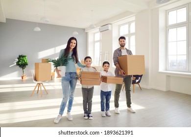 Happy Family Standing In New Home. Smiling Millennial Couple With Kids Carrying Cardboard Boxes Together. Little Boy And Girl Helping Mom And Dad On Moving Day. Buying House, Property, Sale Concept