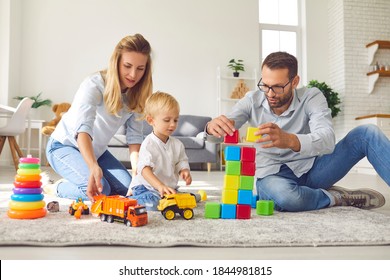 Happy Family Staying At Home And Engaging Their Little Kid In Fun Games. Young Mom, Dad And Child Playing Toy Trucks And Building Tower Of Colorful Blocks Sitting On Warm Floor In The Living-room