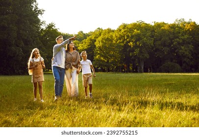Happy family strolling in park in summer, walking on green grass lawn, enjoying beautiful landscapes, looking up, watching planes or birds flying in sky, relaxing and having good time together