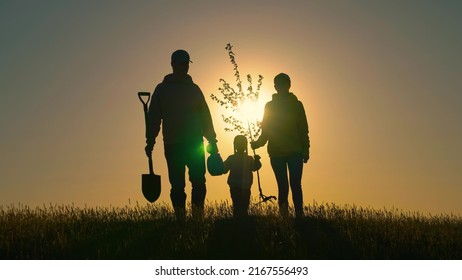 Happy Family Team Planting Tree In Sun Spring Time. Farmer Dad, Mom Child Planting Tree. Silhouette Of Family With Tree At Sunset. Family With Shovel And Watering Can Plants Young Trees Sprout In Soil