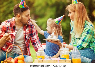 Happy Family Of Three Celebrating Birthday Party In The Park And Eating Cake.