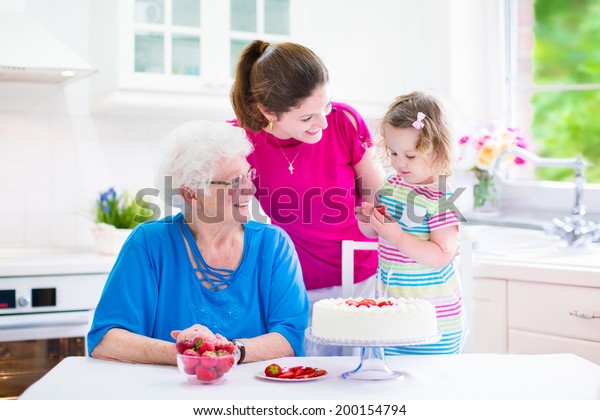 Happy\
family, three generations of women- senior lady, young woman and a\
little toddler girl baking a strawberry cake together in a white\
sunny kitchen with window and modern\
appliances