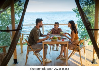 Happy Family Of Three Having Dinner By Wooden Table On Terrace By Seaside