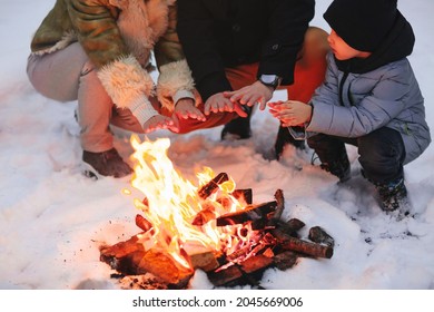 Happy Family Of Three, Mother, Father And Son, In Warm Clothes, Sitting Close To Each Other And Trying To Warm Up By Fire Outside In Cold Snowy Winter Weather, Against Snowbound Forest Background