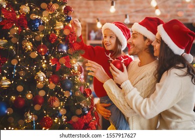 Happy Family Of Three In Red Santa Hats Decorating Xmas Tree With Bubbles And Lights At Home