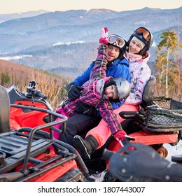 Happy Family Of Three, Wearing Colorful Ski Suits, Spending Great Winter Holiday, Hugging Together In Snowy Mountains On Red Quad Bike, Side View, Ridge Of Mountains On Background.