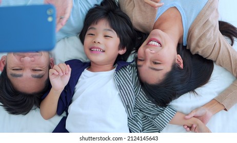 Happy Family Time, Taking Selfie Mobile Phone Of Asian Parents Dad, Mom And Son Laying Together On Bed In Bedroom With Happy, Laughing Smiles. Weekend Parenting With Love Good Time Relaxing At Home.