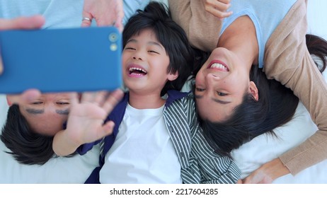 Happy Family Time, Taking Selfie Mobile Phone Of Asian Parents Dad, Mom And Son Laying Together On Bed In Bedroom With Happy, Laughing Smiles. Weekend Parenting With Love Good Time Relaxing At Home.