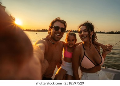 Happy family trio taking a selfie on a boat during sunset. The man is shirtless and wearing sunglasses, the woman is in a bikini top, and the child is in a pink and white outfit. - Powered by Shutterstock