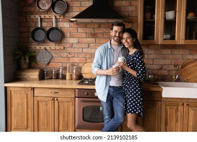 Happy Family Of Two Husband Wife Homeowners Renters Pose For Portrait At New Large Modern Kitchen Embrace With Love Affection Hold Cups Of Tea. Smiling Loving Young Spouses Look At Camera Enjoy Coffee