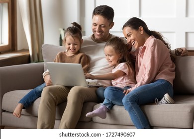 Happy family with two little daughters using laptop together, sitting on cozy sofa at home, smiling father and mother with preschool girls sisters looking at computer screen, shopping online