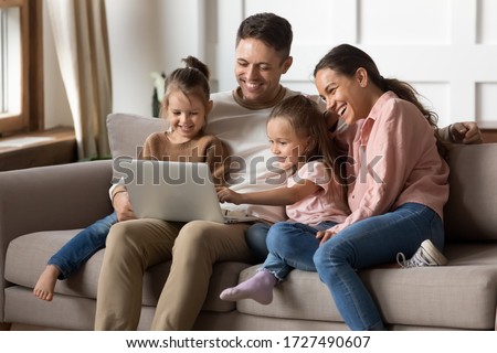 Happy family with two little daughters using laptop together, sitting on cozy sofa at home, smiling father and mother with preschool girls sisters looking at computer screen, shopping online