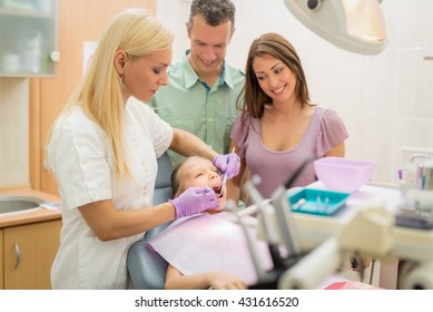 Happy Family At Visit In The Dentist Office. Female Dentist Checking Teeth The Little Girl, Her Parents Standing Next To Her.