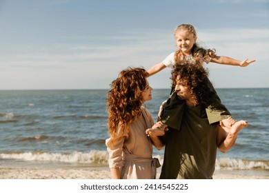 Happy family walking on the beach at sunny day. Parents looking on each other. Spouses holding their little kid piggyback on weekend walking along lake river - Powered by Shutterstock