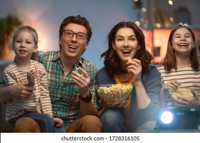 Happy Family Watching Projector, TV, Movies With Popcorn In The Evening At Home. Mother, Father And Daughters Spending Time Together.