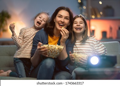 Happy family watching projector, TV, movies with popcorn in the evening at home. Mother and daughters spending time together.