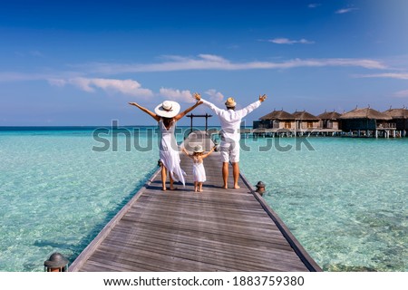 A happy family in white summer clothing on vacation walks along a wooden pier over tropical, turquoise ocean in the Maldives, Indian Ocean