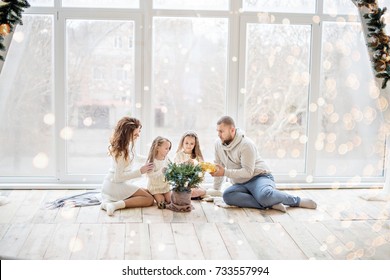 A Happy Family In White Sweaters Prepares To Meet New Year In A Beautiful Loft With Panoramic Windows. Young Parents With Little Daughters Decorate A Small Christmas Tree In A Pot. Copy Space, Bokeh