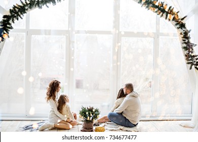 A Happy Family In White Sweaters Prepares To Meet New Year In A Beautiful Loft With Panoramic Windows. Young Parents With Little Daughters Decorate A Small Christmas Tree In A Pot. Copy Space, Bokeh