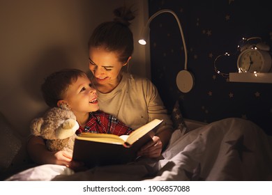 Happy Family Woman And Boy Cuddling And Smiling While Reading Book On Bed Near Lamp At Night At Home