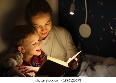 Happy Family Woman And Boy Cuddling And Smiling While Reading Book On Bed Near Lamp At Night At Home