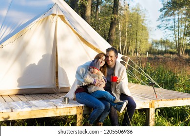 Happy family wraps blanket over themselves while sitting and drinking coffee near canvas tent in the morning in the woods