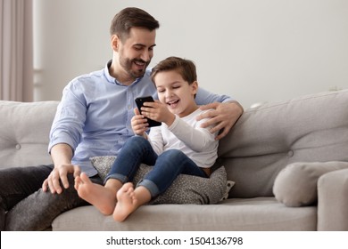 Happy Family Young Dad And Cute Little Kid Child Son Sit On Sofa Relaxing Having Fun With Mobile Phone Playing Games Watching Funny Social Media Videos Laughing Enjoying Using Cellphone At Home