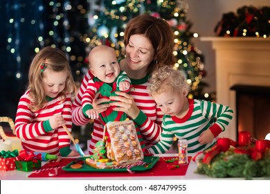 Happy Family, Young Mother With Baby, Little Boy And Girl Making Gingerbread House At Fireplace In Decorated Living Room With Christmas Tree. Baking And Cooking With Children For Xmas At Home.