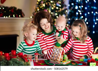 Happy Family, Young Mother With Baby, Little Boy And Girl Making Gingerbread House At Fireplace In Decorated Living Room With Christmas Tree. Baking And Cooking With Children For Xmas At Home.