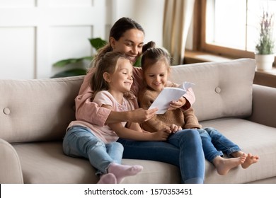 Happy family young mother babysitter hold read book relax embrace cute little children daughters, smiling parent mum tell small kids funny fairy tale story sit on sofa having fun together at home - Shutterstock ID 1570353451