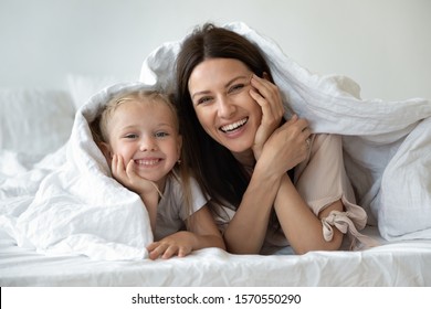 Happy Family Young Mother And Cute Small Kid Daughter Laugh Look At Camera Covered With Warm White Duvet, Smiling Mom Having Fun With Funny Little Child Girl Lay In Cozy Bed Under Blanket, Portrait