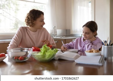 Happy Family Young Mother And Teenage Daughter Studying, Eating Together At Home. Adult Parent Mum Preparing Vegetable Salad Helping Tween School Girl Child Doing Homework Sitting At Kitchen Table.