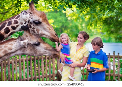 Happy family, young mother with two children, cute laughing toddler girl and a teen age boy feeding giraffe during a trip to a city zoo on a hot summer day