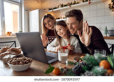 Happy Family Young Parents With Cute Small Kid Child Daughter Holding Xmas Gift Waving Hands Using Laptop Computer At Home Sit At Christmas Table Having Virtual Party Dinner On Video Call Together.