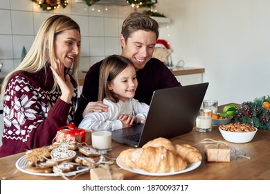 Happy Family Young Parents With Cute Small Kid Child Daughter Waving Hands Using Laptop Computer At Home Sitting At Christmas Table Having Virtual Party Dinner On Video Conference Call Together.