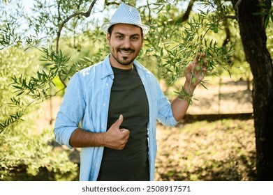 Happy farmer in a hat smiles and gives a thumbs up while inspecting an olive tree in a sunny grove, exuding positivity and confidence in nature - Powered by Shutterstock